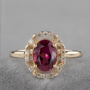 Peut inclure: Une bague en or ornée d'une grosse pierre rouge foncé de forme ovale. La pierre précieuse est entourée de petites pierres claires et sertie dans un sertissage en or décoratif. La bague a un anneau délicat.