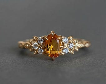 Puede incluir: Un anillo de oro con una piedra preciosa de citrino naranja de corte ovalado en el centro. La piedra está flanqueada por grupos de pequeñas piedras blancas redondas y transparentes engastadas en la banda de oro con un diseño metálico texturizado.
