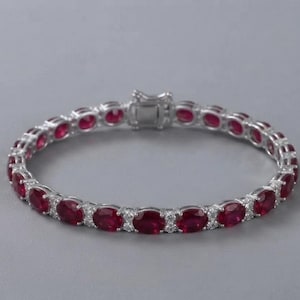 May include: A silver-toned tennis bracelet featuring a series of oval-cut deep red rubies alternating with small, clear, round-cut accent stones. The bracelet is secured with a rectangular metal box clasp mechanism at the top center.