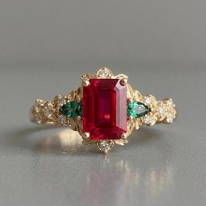 May include: A gold ring featuring a large, rectangular, deep red gemstone as the focal point. Two smaller, pear-shaped green gems flank the red stone. The band is adorned with small diamonds and decorative filigree.