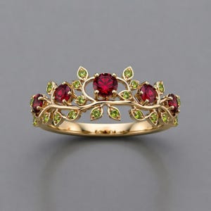May include: A gold-colored ring featuring a floral design. The ring has a series of red gemstones and green leaf accents. The gemstones are round and set in a delicate, nature-inspired pattern. The ring is set against a gray background.