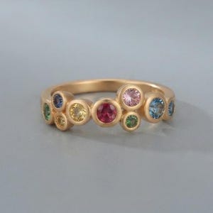 Può includere: Un anello in oro con un gruppo di pietre preziose rotonde incastonate a lunetta in vari colori, tra cui rosa, blu, giallo e verde. La fascia ha una finitura opaca liscia e le pietre sono disposte in un motivo asimmetrico a forma di bolle.