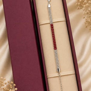 May include: A silver-toned bracelet featuring a central row of rectangular red stones flanked by two rows of clear, sparkling crystals. The bracelet includes a lobster claw clasp and an adjustable chain extension, presented inside a deep burgundy box.