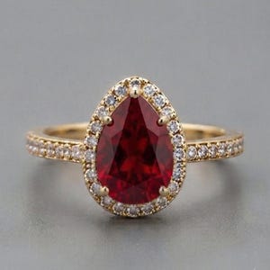 Könnte beinhalten: Ein Goldring mit einem großen, birnenförmigen, tiefroten Edelstein in der Mitte. Der zentrale Stein ist von einem Kranz aus kleinen, klaren, rund geschliffenen Steinen umgeben. Die Ringschiene ist ebenfalls mit einer Reihe kleiner, klarer Steine besetzt.