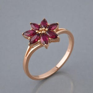 Op de afbeelding: Een roségouden ring met een bloemdesign. De bloem bestaat uit zeven kastanjebruine, marquise-geslepen edelstenen. De ring heeft een split-schacht ontwerp en is geplaatst tegen een grijze achtergrond.