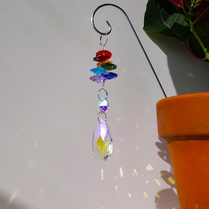 Op de afbeelding: Een decoratieve kristallen zonnevanger met een regenboog van gekleurde glazen kralen en een groot druppelvormig kristal. De zonnevanger hangt aan een metalen haak en werpt kleurrijke lichtpatronen. Een terracotta pot met een plant staat op de achtergrond.