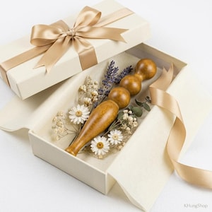 Puede incluir: Una herramienta de masaje de madera con un mango redondeado y tres cabezas esféricas, presentada en una caja de regalo blanquecina con una cinta dorada. La caja está forrada con papel y contiene flores secas y ramitas de lavanda.