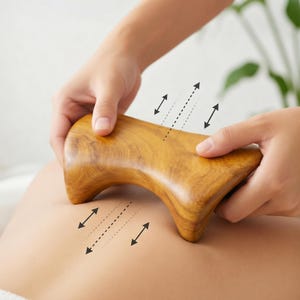 May include: A wooden massage tool with a curved design is held over a person's back. The tool is light brown with a smooth finish. Arrows indicate the direction of use. The tool is designed for massage therapy and relaxation.