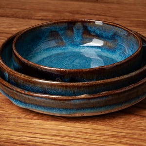 Op de afbeelding: Twee ronde keramische kommen die op elkaar zijn gestapeld. De kommen hebben een levendig blauw geglazuurde binnenkant die overgaat in een donkerbruine, aardse rand. De buitenoppervlakken vertonen een gevlekte textuur met zichtbare blauwe en bruine tinten.