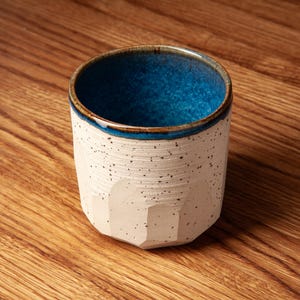 Può includere: Una tazza in ceramica con esterno bianco sporco maculato e interno smaltato in blu vibrante. Il recipiente presenta una parte inferiore sfaccettata con distinti piani verticali e un bordo liscio e arrotondato. La superficie è opaca.