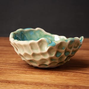 Op de afbeelding: Een kleine, ronde keramische kom met een textuur die lijkt op een honingraat aan de buitenkant. De buitenkant is crèmekleurig, terwijl de binnenkant is voorzien van een glanzend, levendig turquoise glazuur dat zich in de holtes verzamelt.