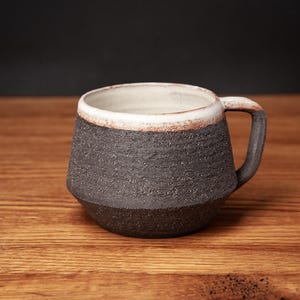 May include: A ceramic mug with a dark charcoal, textured exterior and a smooth, off-white interior. The rim features a subtle, rustic reddish-brown accent where the two textures meet, and the mug includes a single, matching dark handle.