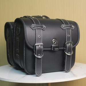 May include: Black leather motorcycle saddlebags with white stitching. The bags have silver buckles, a lock, and a classic design. The saddlebags are sitting on a white surface.