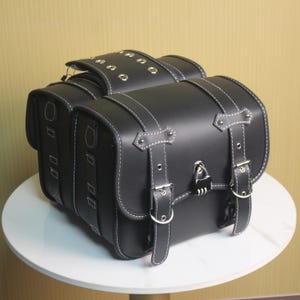 May include: Black motorcycle saddlebags with white stitching and silver buckles. The bags have a rectangular shape with rounded corners and a lock on the front. The top flap has silver grommets.