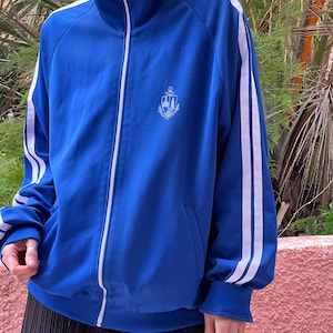 Puede incluir: Chaqueta deportiva azul real con rayas blancas en las mangas y cremallera blanca. La chaqueta tiene cuello alto y un pequeño emblema bordado en el pecho. Estilo deportivo.
