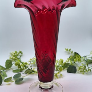 May include: A tall, deep red glass vase featuring a flared, ruffled rim and a subtle spiral pattern throughout the body. The vase stands on a clear, circular glass base, showcasing a translucent finish that catches the light.