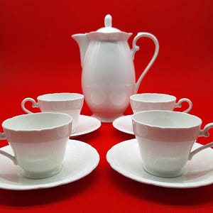 May include: A white porcelain tea set comprising a tall teapot with a lid and handle, alongside four matching teacups and saucers. Every item features a smooth, glossy finish with scalloped edges along the rims of the cups and the matching saucers.