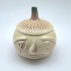 May include: A ceramic onion-shaped container with a lid. The lid is tan with the word "ONIONS" and a green stem. The container has a face with a sad expression and teardrops. The container is off-white.