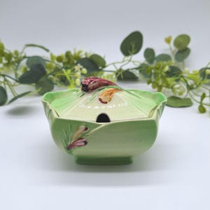 May include: A light green ceramic sugar bowl with a matching lid featuring a small cutout for a spoon. The bowl and lid are decorated with embossed and painted floral motifs in shades of yellow, purple, and green, resembling cabbage leaves.