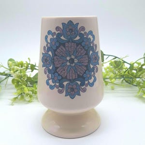 May include: A cream-coloured ceramic vase with a pedestal base, featuring a central circular floral motif in shades of blue, purple, and dark grey. The design consists of symmetrical petals and leaves arranged in a repeating pattern on the front.