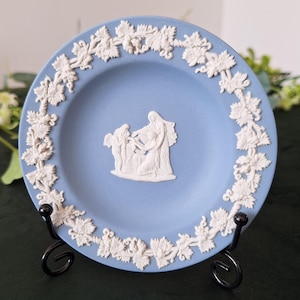 May include: A pale blue decorative plate with a white relief design. The plate features a central scene of figures and a border of grape leaves and vines. The plate is displayed on a black metal stand.