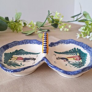 May include: A two-section ceramic serving dish with a handle. The dish has a white base with a blue scalloped edge and a painted design featuring figures and foliage. The handle is yellow and blue.