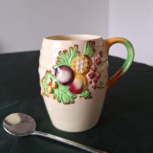 May include: Cream-colored ceramic mug with a fruit design, including plums, grapes, and peaches. The handle transitions from brown to green. A silver spoon rests in front of the mug. The mug has a textured barrel-like design.