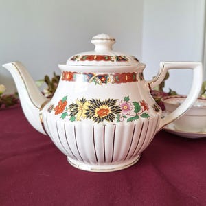 May include: A white ceramic teapot with a lid, decorated with vibrant floral patterns in orange, yellow, and pink. The teapot features gold trim and vertical lines. A matching teacup is visible.