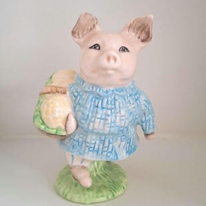 May include: A ceramic pig figurine wearing a blue sweater and carrying a green and yellow bundle. The pig stands on a green grass base. The pig has pink skin, brown ears, and black eyes.