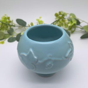 May include: A light blue ceramic bowl with a rounded, spherical shape and a smooth finish. The exterior surface features a raised, embossed pattern of ivy leaves and winding vines that wrap around the circumference of the vessel.