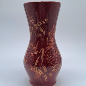 May include: A burgundy vase with a speckled finish. The vase has a flared top and a rounded body. Gold botanical designs, including wheat and leaves, are painted on the surface. The vase is a decorative item.