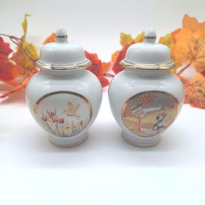 May include: Two white porcelain ginger jars with lids, each featuring a gold-rimmed circular medallion. One jar displays a hummingbird among flowers, while the other depicts a figure in a landscape with a structure, both accented with gold trim.