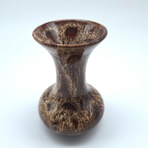 May include: A small, brown ceramic vase with a speckled pattern. The vase has a rounded base, a slender neck, and a flared rim. The color palette includes shades of brown and cream, creating a textured appearance. The vase is likely for decorative purposes.