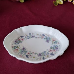 May include: White ceramic oval dish with a scalloped edge. The dish is decorated with a floral pattern in shades of pink, blue, and purple. The flowers are arranged in a circular pattern in the center of the dish.