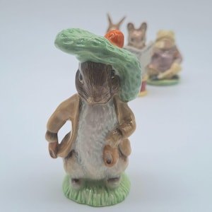 May include: A ceramic Peter Rabbit figurine, wearing a green hat with an orange detail and a tan jacket. The rabbit stands on a patch of green grass. Other figurines are slightly out of focus in the background.