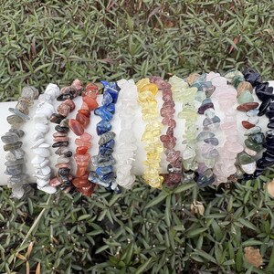 Può includere: Una collezione di braccialetti con schegge di pietre preziose in vari colori, tra cui grigio, bianco, rosso, blu, giallo e rosa. I braccialetti sono infilati e presentati su una superficie cilindrica bianca, su uno sfondo di fogliame verde.