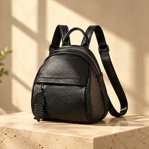 Könnte beinhalten: Ein kleiner schwarzer Rucksack mit genarbter Textur, ausgestattet mit einem abgerundeten Hauptfach, einer vorderen Reißverschlusstasche und verstellbaren Schultergurten. Ein dekorativer schwarzer Fischgrätenanhänger hängt am Reißverschluss.