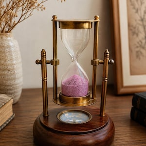 May include: A rotating hourglass featuring a brass frame and clear glass bulbs filled with vibrant purple sand. The hourglass is mounted on a circular dark wood base that incorporates a functional compass embedded in the center of the platform.
