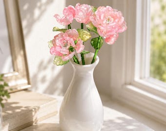 May include: A white ceramic vase with a narrow neck holds a bouquet of translucent pink artificial flowers with green stems and leaves. The flowers have layered petals and a glossy finish, creating a delicate appearance in this decorative arrangement.