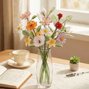 May include: A colorful glass flower arrangement in a clear vase. The bouquet includes orange poppies, pink and white daisies, and other assorted flowers. A teacup and saucer, a small succulent, and an open book are also on the table.