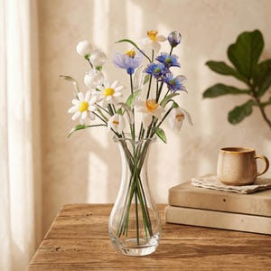 May include: A clear glass vase holds a bouquet of artificial flowers. The flowers include white daisies with yellow centers, blue and purple blooms, and white bell-shaped flowers. The vase sits on a wooden table, with a coffee cup and books in the background.