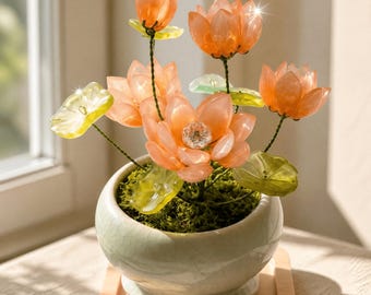 May include: A decorative arrangement featuring translucent orange lotus flowers with a central crystal accent, paired with light green leaves on thin stems. The flowers are planted in a round, pale green ceramic pot with a crackle glaze finish.