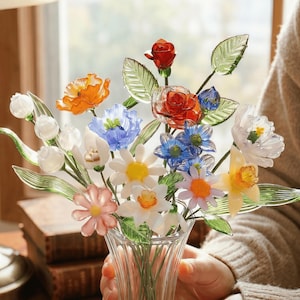 May include: A colorful glass flower bouquet in a clear vase. The arrangement includes orange poppies, red roses, white daisies, and blue flowers. The glass flowers have green leaves and are held in a clear, fluted vase.