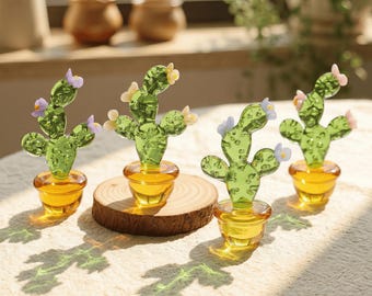 May include: Four decorative glass cactus figurines with yellow pots and colorful flowers. The cacti are green with small, delicate flowers in shades of pink, yellow, and purple. The pots are a warm amber color, and one cactus sits on a wooden slice.