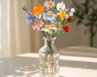 May include: A clear glass vase with a bouquet of colorful, handcrafted flowers. The flowers include orange, blue, white, and pink blooms. The vase has the text "Mom's Garden" and names of people on it.