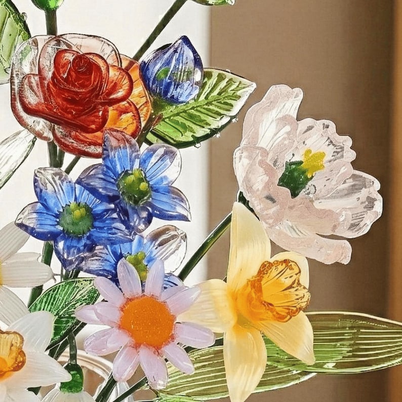 May include: A close-up of a glass floral arrangement featuring a red rose, blue and white daisies, and a yellow daffodil. The flowers are made of translucent glass with green stems and leaves. The arrangement is set against a neutral background.