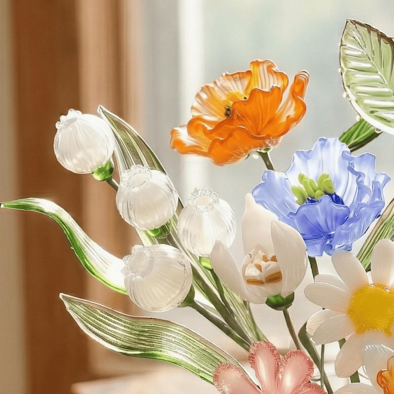 May include: A colorful glass flower arrangement featuring orange poppies, blue and white flowers, and white buds. The glass flowers have intricate details and are set against a blurred background.
