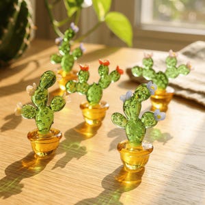 May include: Several glass cactus figurines in small, gold-colored pots are arranged on a wooden surface. The cacti are green with small, colorful flower accents. The pots have a glossy finish, and the scene is well-lit.