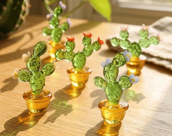 May include: Several glass cactus figurines in small, gold-colored pots are arranged on a wooden surface. The cacti are green with small, colorful flower accents. The pots have a glossy finish, and the scene is well-lit.