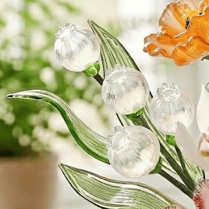 May include: Close-up of a glass floral arrangement featuring clear, spherical flower buds with textured details. The arrangement includes green leaves and a vibrant orange flower, set against a blurred background, creating a delicate and artistic display.